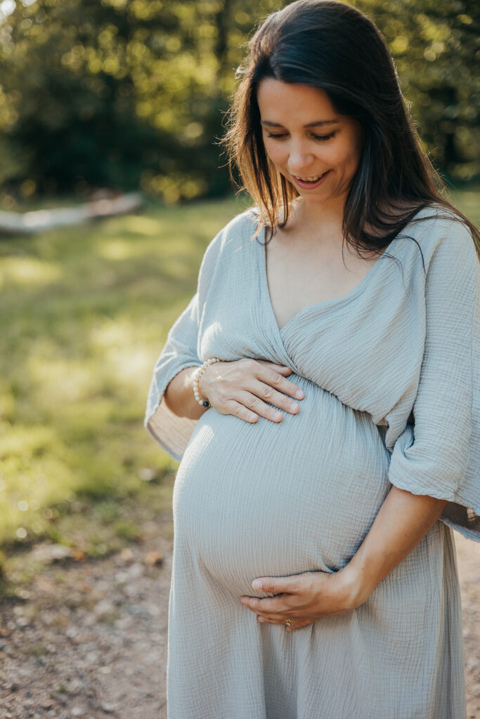 Glücklich lächelnde Schwangere betrachtet ihren Babybauch bei einem Fotoshooting im Grünen