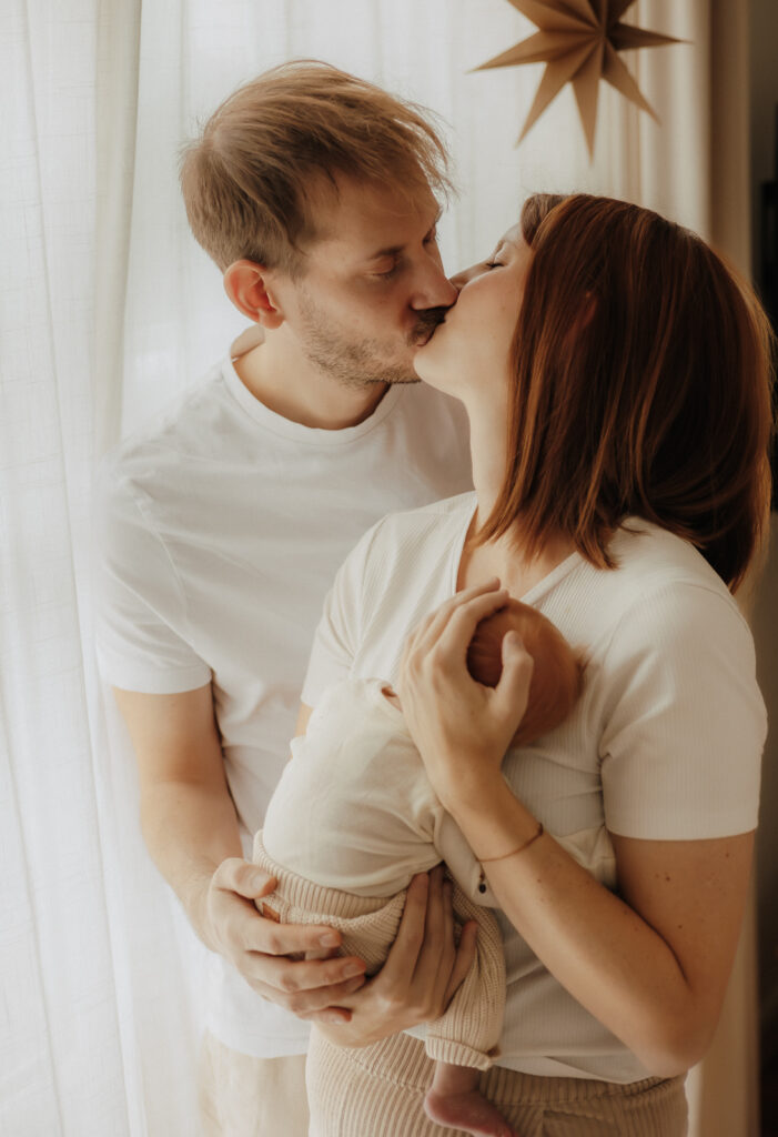 Eltern küssen sich zärtlich, während sie ihr Neugeborenes im Arm halten – Familienfotografin Saarland