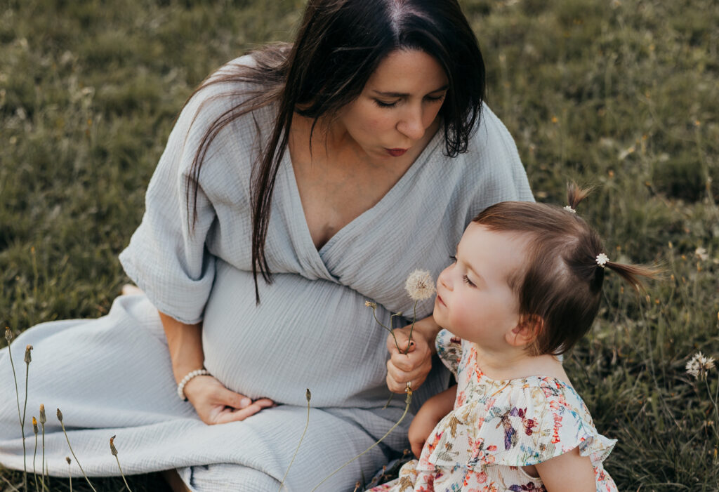 Schwangere Mutter sitzt mit ihrem Kleinkind auf einer Wiese und beobachtet es beim Pusten einer Pusteblume