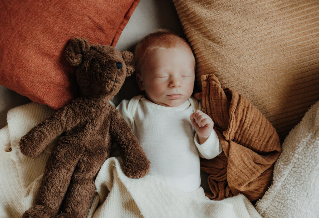 Schlafendes Neugeborenes liegt zwischen weichen Kissen neben einem Teddybären