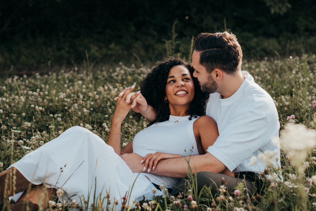 Paar liegt entspannt in einer Blumenwiese, schaut sich verliebt an und lacht gemeinsam