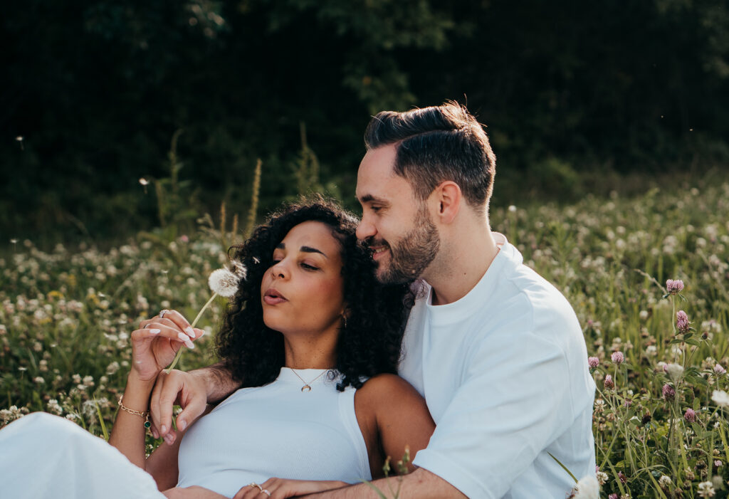 Paar liegt eng beieinander in einer sommerlichen Wiese und pustet eine Pusteblume.